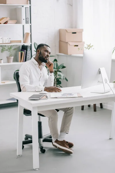 Pensativo guapo afroamericano hombre de negocios sentado en la mesa y mirando hacia arriba en la oficina - foto de stock