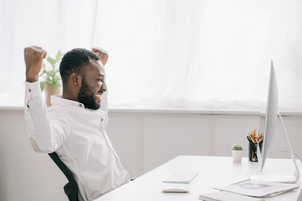 Боковой вид счастливого африканского бизнесмена, работающего за компьютером в офисе и показывающего жест 