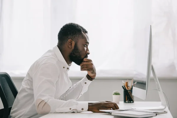 Vista laterale del bell'uomo d'affari afroamericano che riposa sul mento a portata di mano e lavora al computer in ufficio — Foto stock