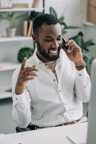 Cheerful handsome african american businessman talking by smartphone and gesturing in office — Stock Photo