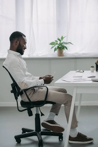 Seitenansicht eines gut aussehenden afrikanisch-amerikanischen Geschäftsmannes, der am Tisch sitzt und sein Smartphone im Büro benutzt — Stockfoto