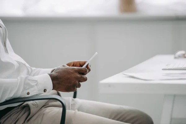 Обрезанный образ африканского американского бизнесмена, сидящего на стуле и использующего смартфон в офисе — стоковое фото