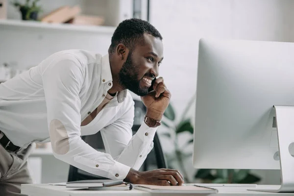 Vista lateral del guapo hombre de negocios afroamericano hablando por teléfono inteligente y utilizando la computadora en la oficina — Stock Photo
