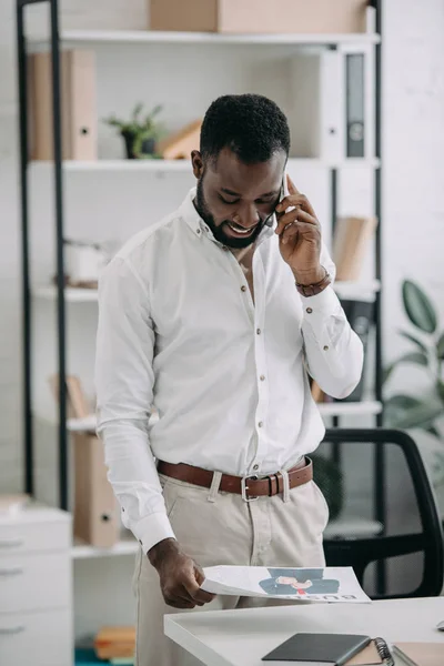 Apuesto hombre de negocios afroamericano hablando por teléfono inteligente en la oficina y leyendo el periódico - foto de stock