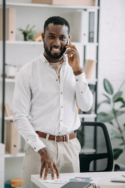 Apuesto hombre de negocios afroamericano de pie cerca de la mesa y hablando por teléfono inteligente en la oficina - foto de stock