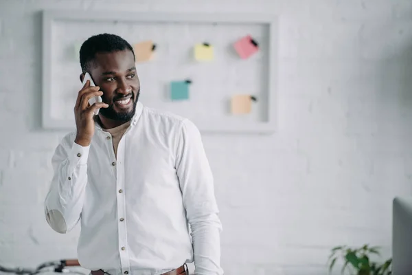 Alegre guapo africano americano hombre de negocios hablando por teléfono inteligente en la oficina y mirando hacia otro lado - foto de stock