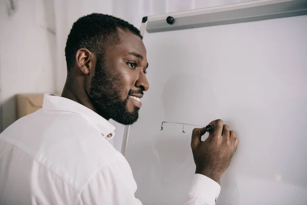 Homme d'affaires afro-américain souriant dessinant sur tableau à feuilles mobiles au bureau et détournant les yeux — Photo de stock