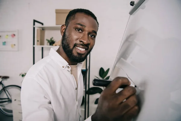Улыбающийся красивый африканский бизнесмен, рисующий на флипчарте в офисе и смотрящий в камеру — стоковое фото