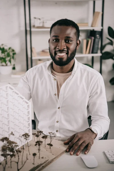 Усміхнений афроамериканський архітектор сидить біля архітектурної моделі на столі в офісі і дивиться на камеру — стокове фото