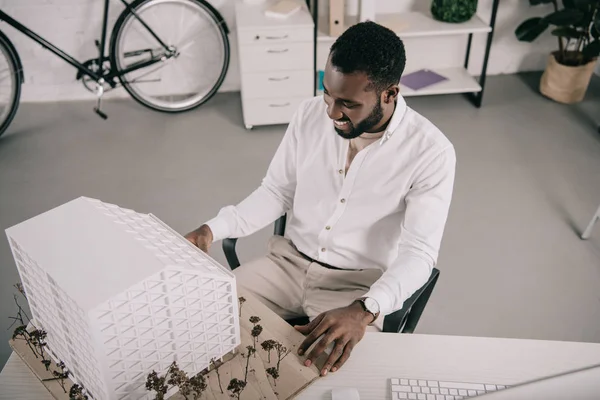 Високий кут зору веселого афроамериканського архітектора, який дивиться на архітектурну модель в офісі — стокове фото