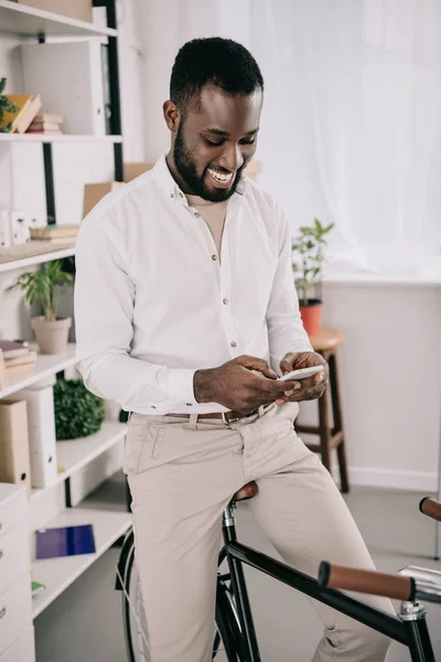 Fröhlich gut aussehender afrikanisch-amerikanischer Geschäftsmann sitzt auf dem Fahrrad und benutzt Smartphone im Büro — Stockfoto