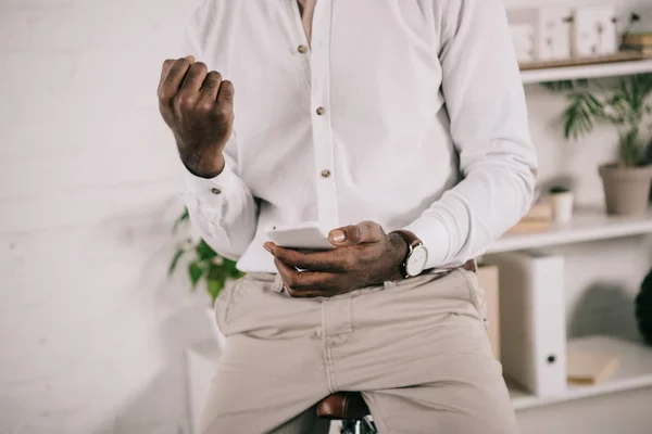 Imagen recortada de hombre de negocios afroamericano sentado en bicicleta, utilizando el teléfono inteligente y mostrando sí gesto en la oficina — Stock Photo