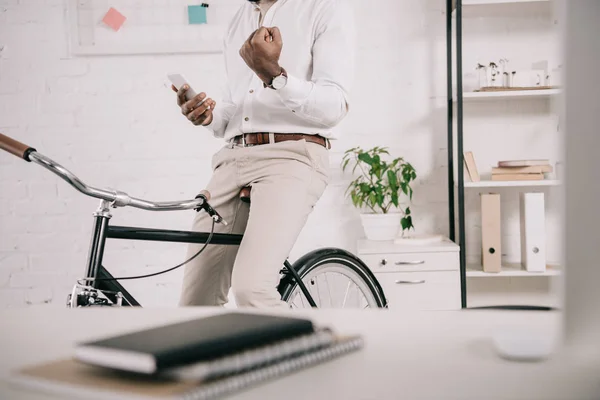 Imagen recortada de hombre de negocios afroamericano sentado en bicicleta, utilizando el teléfono inteligente y mostrando sí gesto en la oficina - foto de stock