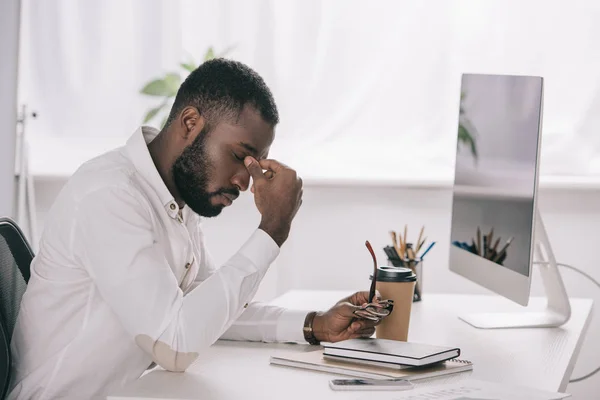 Side view of tired african american businessman touching nose bridge at table with computer in office — Stock Photo