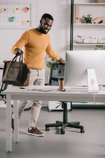 Lächelnd gutaussehender afrikanisch-amerikanischer Designer in orangefarbenem Pullover mit Aktentasche und Blick auf den Computer im Büro — Stockfoto