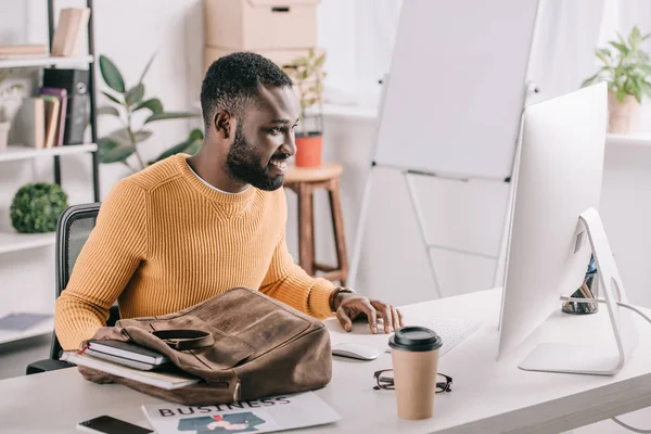 Apuesto diseñador afroamericano en suéter naranja tomando cuadernos de maletín y mirando la computadora en la oficina — Stock Photo