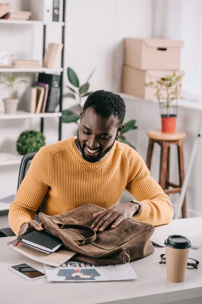 Guapo diseñador afroamericano en suéter naranja tomando cuadernos de maletín en la oficina - foto de stock