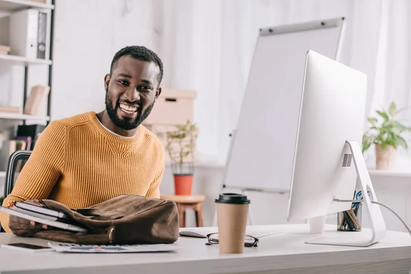 Lächelnd gutaussehender afrikanisch-amerikanischer Designer in orangefarbenem Pullover, der im Büro Notizbücher aus der Aktentasche nimmt — Stockfoto