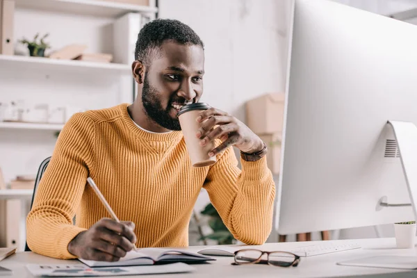 Handsome african american designer in orange sweater drinking coffee and drawing in notebook in office — Stock Photo