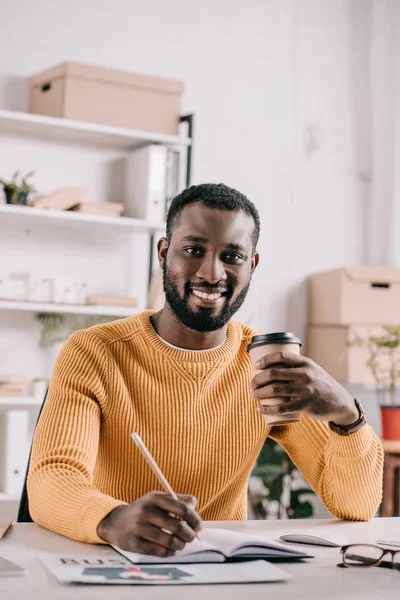 Handsome african american designer drawing in notebook and holding coffee in paper cup in office — Stock Photo