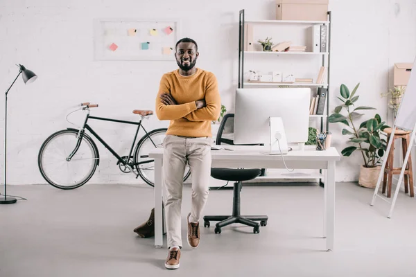 Belo designer afro-americano com braços cruzados apoiados na mesa e olhando para a câmera no escritório — Fotografia de Stock