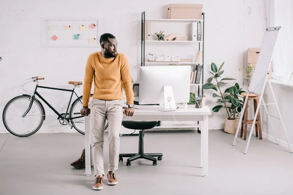 Красивый африканский американский дизайнер в оранжевом свитере, опирающийся на стол и смотрящий в сторону в офисе — стоковое фото