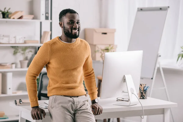 Alegre bonito designer afro-americano em camisola laranja inclinado na mesa e olhando para longe no escritório — Fotografia de Stock