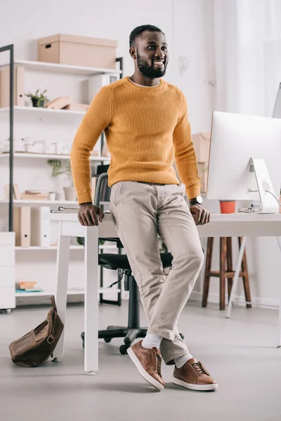 Lächelnd gutaussehender afrikanisch-amerikanischer Designer in orangefarbenem Pullover, der sich auf den Tisch lehnt und im Büro wegschaut — Stockfoto