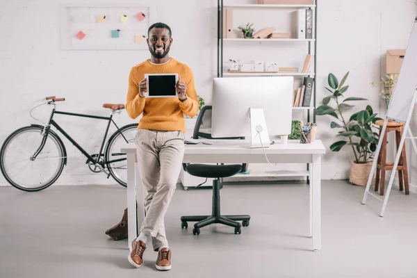 Alegre guapo africano americano diseñador en suéter naranja mostrando tableta con pantalla en blanco en la oficina moderna - foto de stock