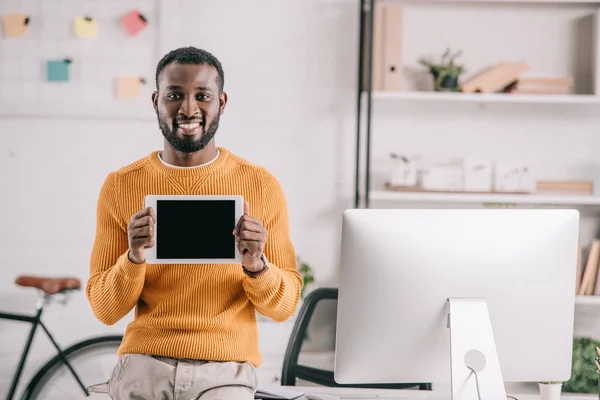 Sonriente guapo africano americano diseñador en naranja suéter mostrando tableta con pantalla en blanco en la oficina - foto de stock