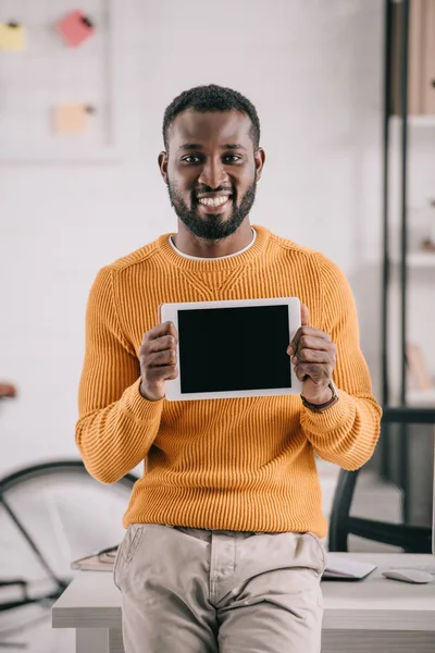 Retrato del guapo diseñador afroamericano sonriente en suéter naranja mostrando tableta con pantalla en blanco en la oficina - foto de stock
