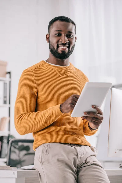 Sonriente guapo africano americano diseñador en naranja suéter celebración tableta y mirando a la cámara en la oficina - foto de stock