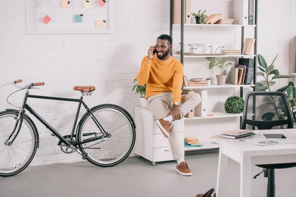 Sonriente guapo africano americano diseñador en naranja suéter hablando por teléfono inteligente en la oficina - foto de stock