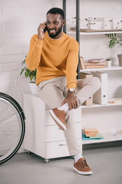 Schöner afrikanisch-amerikanischer Designer in orangefarbenem Pullover, der im Büro per Smartphone spricht — Stockfoto