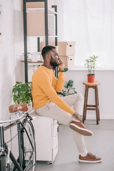 Afrikanischer amerikanischer Designer im gelben Pullover spricht im modernen Büro mit Fahrrad auf dem Smartphone — Stockfoto