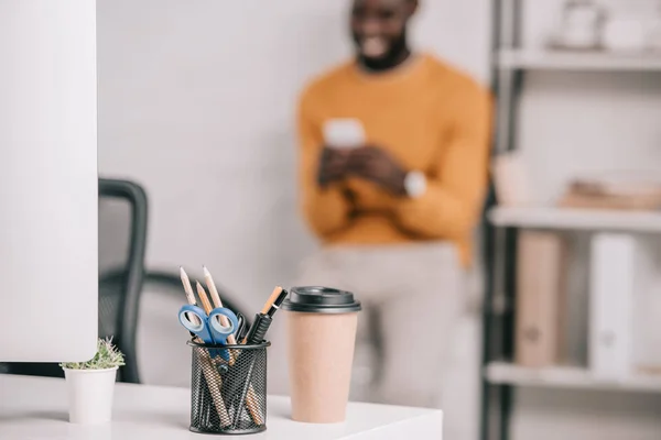 Selective focus of computer, coffee to go and office supplies on workplace and designer standing in modern office — Stock Photo