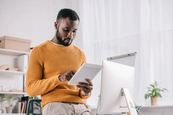 Концентрований афроамериканський дизайнер в жовтому светрі, що працює з цифровим планшетом в сучасному офісі — стокове фото