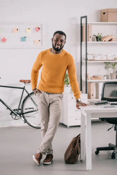 Щасливий афроамериканський дизайнер в жовтому светрі, що стоїть в сучасному офісі — стокове фото