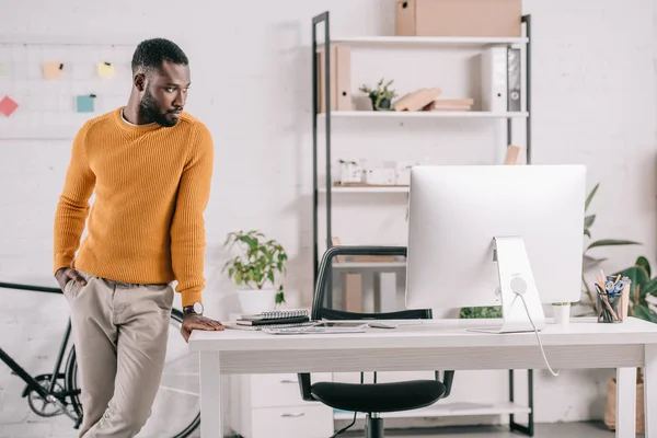 Gerente afroamericano en suéter amarillo de pie en la oficina moderna con computadora - foto de stock