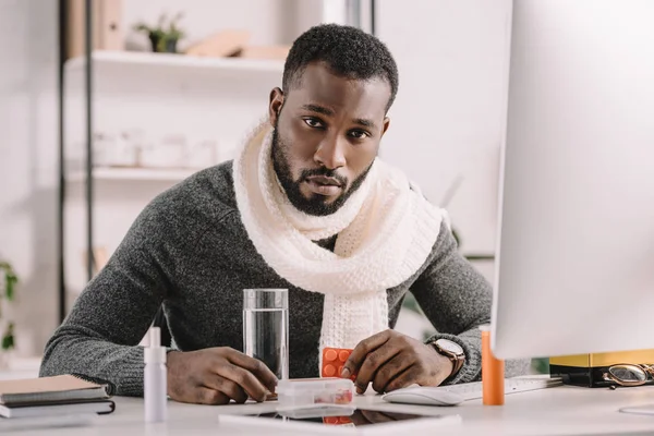 Homme d'affaires afro-américain malade avec des pilules et un verre d'eau assis sur le lieu de travail au bureau — Photo de stock