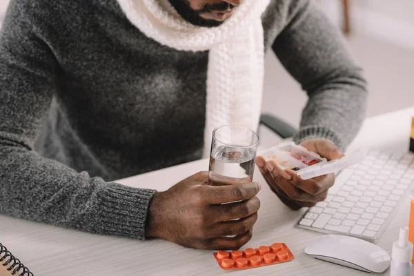Cropped view of ill man with pills and glass of water sitting at workplace — Stock Photo