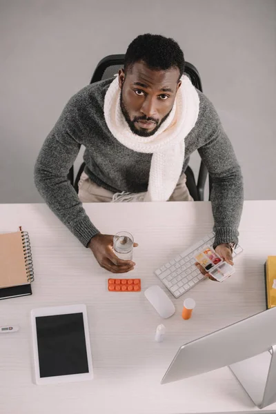 Vue aérienne d'un homme d'affaires afro-américain malade avec des pilules et un verre d'eau assis dans un espace de travail — Photo de stock