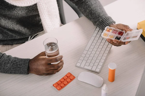 Cropped view of diseased businessman with pills and glass of water sitting at workplace — Stock Photo