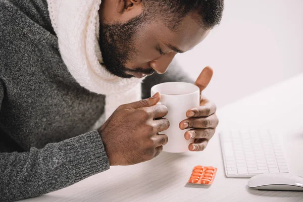 Втомлений хворий афроамериканський чоловік з чашкою гарячого напою та таблетками, що сидять на робочому місці — стокове фото