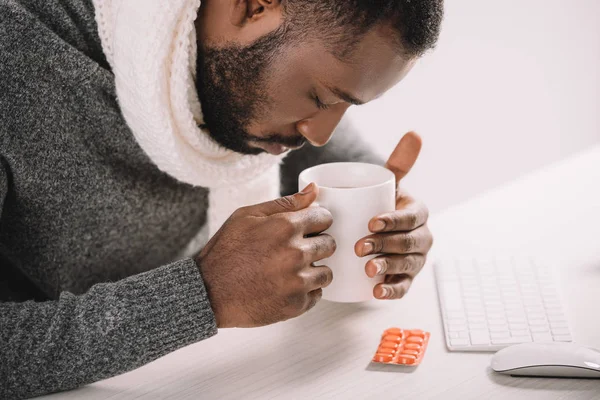 Fatigué malade afro-américain homme avec tasse de boisson chaude et des pilules assis sur le lieu de travail — Photo de stock