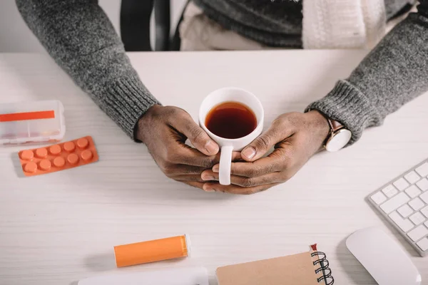 Vue recadrée de l'homme malade avec une tasse de thé chaud et des médicaments assis sur le lieu de travail — Photo de stock