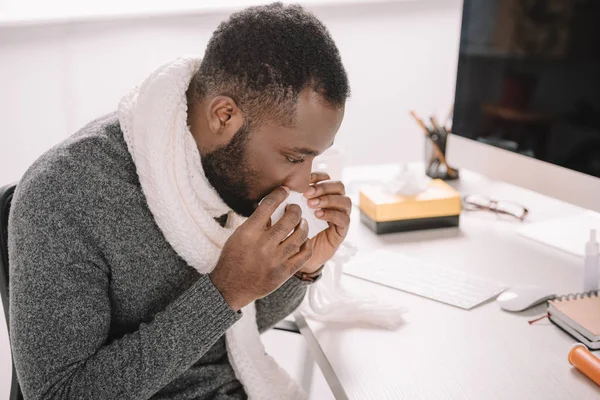 Ill african american man with runny nose holding napkin while sitting at workplace — Stock Photo