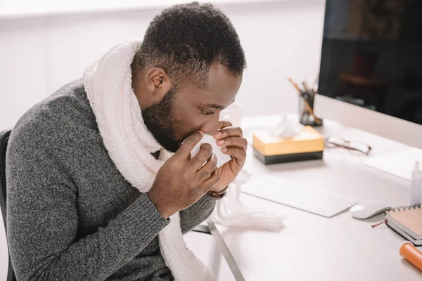 Ill africano americano uomo con naso che cola tenendo tovagliolo mentre seduto sul posto di lavoro — Foto stock