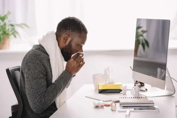 Homme afro-américain malade avec nez qui coule tenant la serviette tout en étant assis sur le lieu de travail avec ordinateur — Photo de stock
