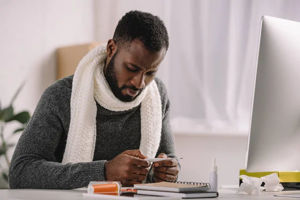 Homme afro-américain malade regardant thermomètre électronique et assis au bureau avec des médicaments — Photo de stock